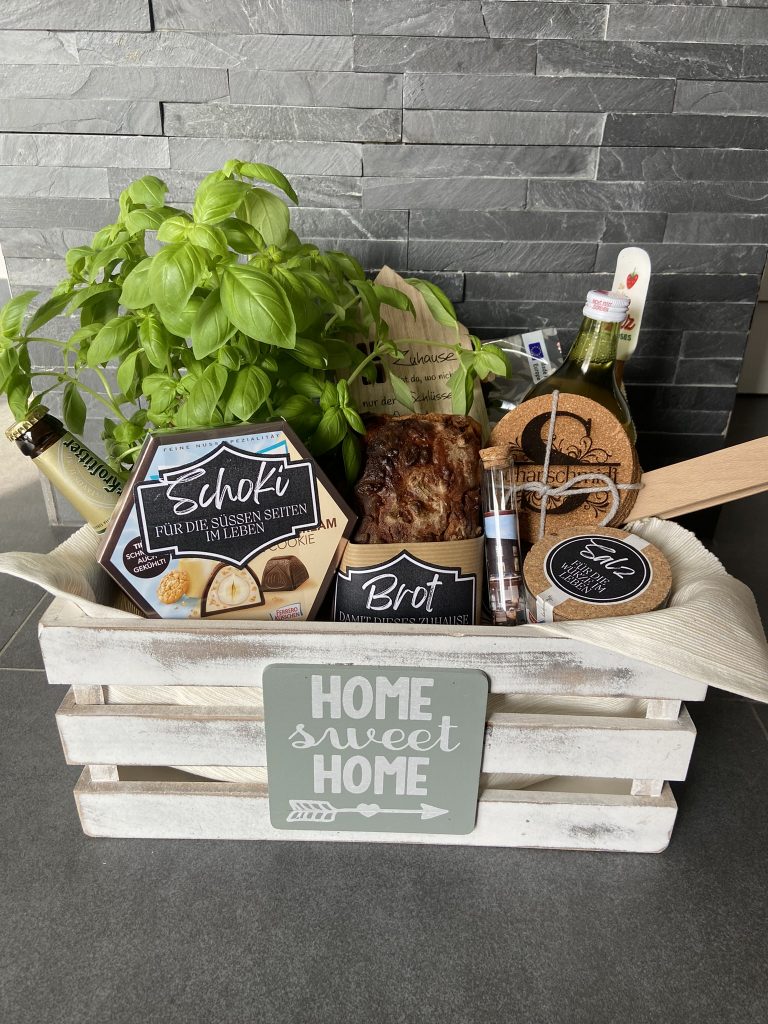 Geschenkbox „Home Sweet Home“ mit Basilikum, Brot, Süßigkeiten, Getränken und gravierten Accessoires aus Holz und Kork.