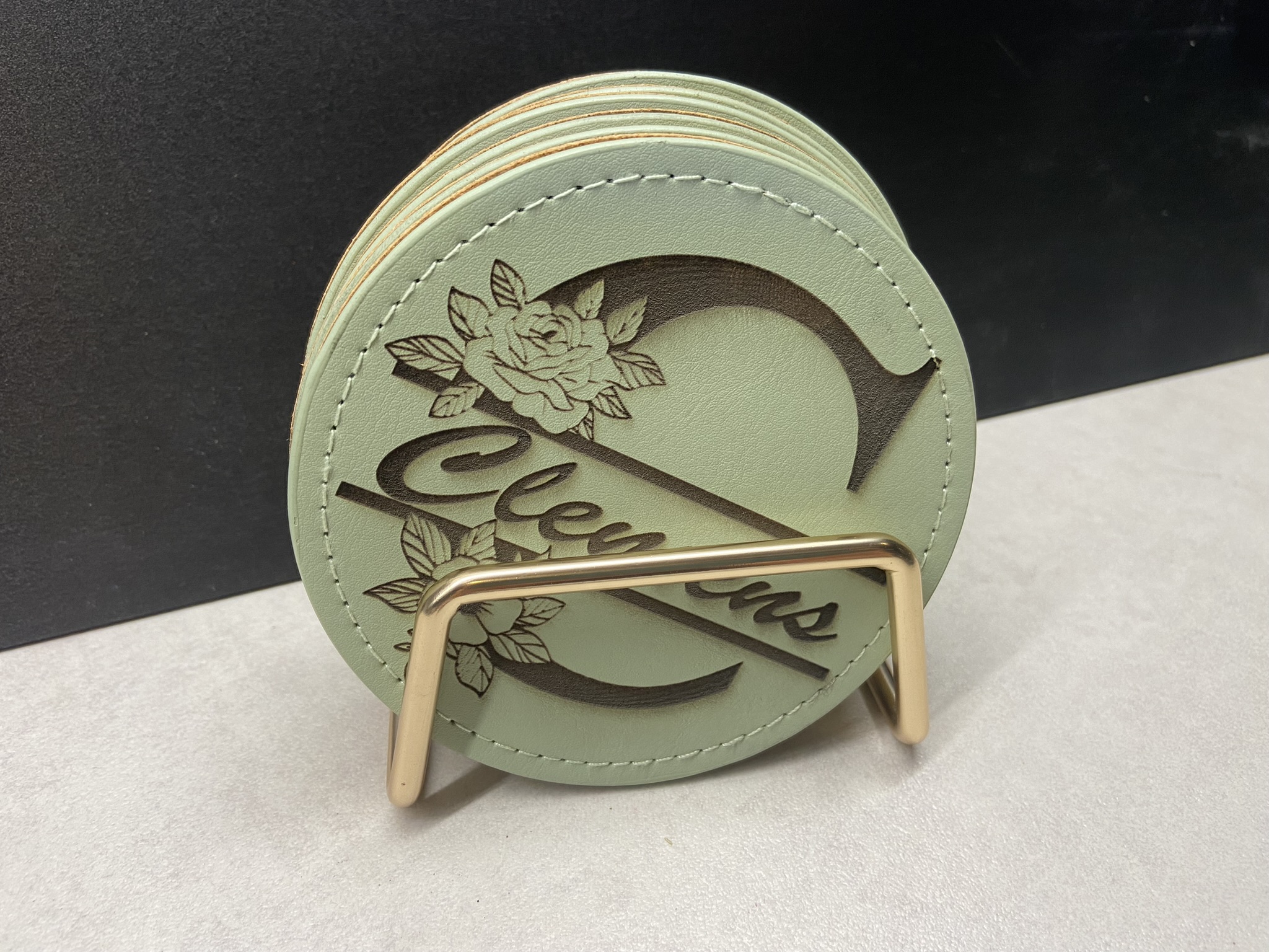 Personalisierte Lederuntersetzer mit Gravur Runde Lederuntersetzer mit Namensgravur „Clemens“ und floralem Design im goldfarbenen Ständer.